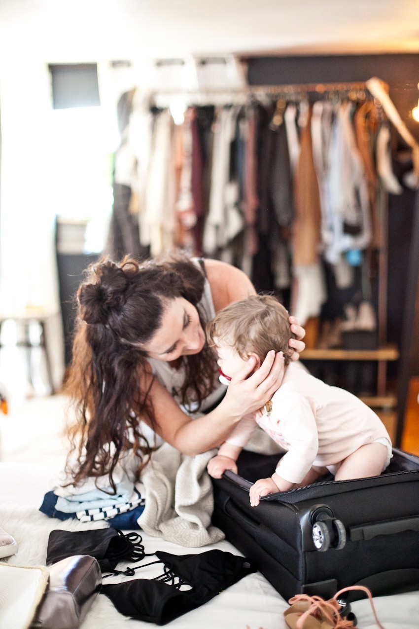 Summer travel and beach getaways, how to prepare and pack light. Get that summer glow and get ready to enjoy a beach escape with these tips and tricks! | Beach Vacation by popular Florida travel blog, Fresh Mommy Blog: image of a woman wearing a white tank top and black leggings and packing her black suitcase that her baby is sitting in. 