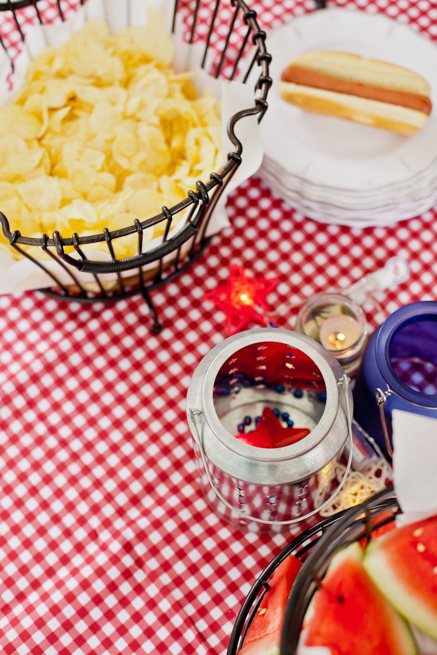 Red White and Blue fun, and a DIY Americana Hot Dog Bar! Set up toppings and flavors for all kinds of hot dogs from around the U.S. Chicago style, Boston, Detroit, Philly, Carolina hot dog and more! |  Hot Dog Bar by popular Florida lifestyle blog, Fresh Mommy Blog: image of a black wire basket filled with potato chips next to a stack of what plates on a red and white gingham table cloth. 