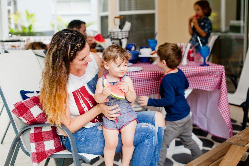 Red White and Blue fun, and a DIY Americana Hot Dog Bar! Set up toppings and flavors for all kinds of hot dogs from around the U.S. Chicago style, Boston, Detroit, Philly, Carolina hot dog and more! |  Hot Dog Bar by popular Florida lifestyle blog, Fresh Mommy Blog: image of a mom sitting outside and hold her baby as they eat a slice of watermelon. 