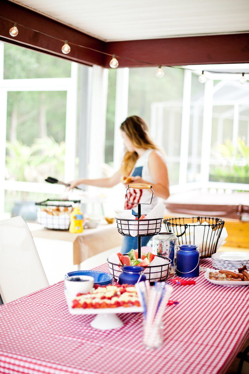Red White and Blue fun, and a DIY Americana Hot Dog Bar! Set up toppings and flavors for all kinds of hot dogs from around the U.S. Chicago style, Boston, Detroit, Philly, Carolina hot dog and more! |  Hot Dog Bar by popular Florida lifestyle blog, Fresh Mommy Blog: image of a woman wearing an American flag tank top and assembling a hot dog at a table with a red and white check table cloth, tiered black wire serving dish with watermelon slices, white serving plate of hot dogs, and American flag fruit platter. 