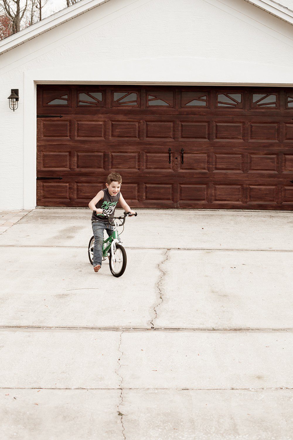 Tips for a DIY Garage Door Makeover and how to Gel Stain a Garage Door to Look Like Wood by popular lifestyle blogger Tabitha Blue of Fresh Mommy Blog: image of young boy riding a bike in front of a white house with a faux wood garage door. 