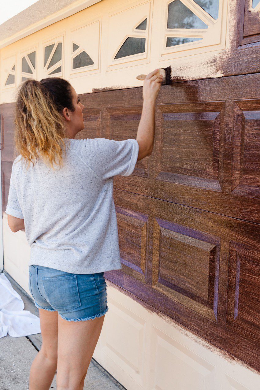 Transform your garage door to a gorgeous faux wood door with this easy gel stain DIY garage door makeover by popular diy blogger Tabitha Blue of Fresh Mommy Blog: image of woman painting a cream garage door to look like faux wood.