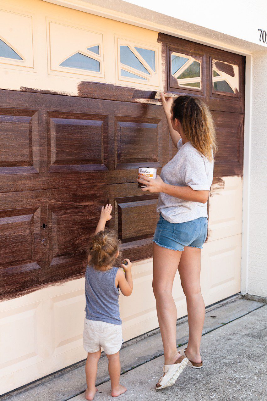 Tips for a DIY Garage Door Makeover and how to Gel Stain a Garage Door to Look Like Wood by popular lifestyle blogger Tabitha Blue of Fresh Mommy Blog: image of woman and young girl painting cream garage door to look like faux wood