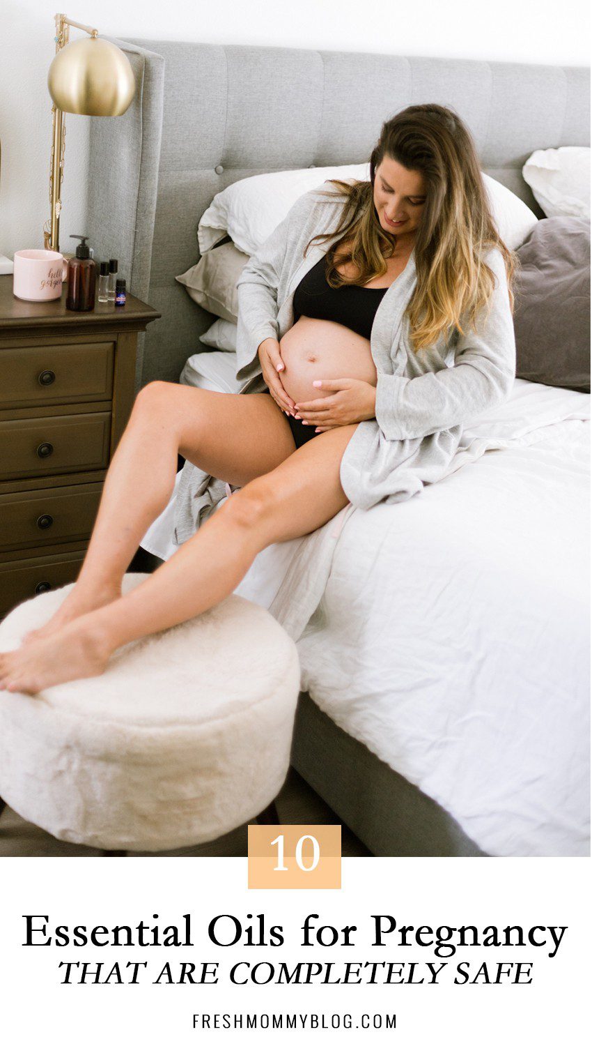 Top 10 Essential Oils While Pregnant That are Completely Safe by popular Florida lifestyle blog, Fresh Mommy: image of woman sitting on her bed in a grey robe, black sports bra, and black underwear and looking down at her pregnant belly while she places her hand on it.