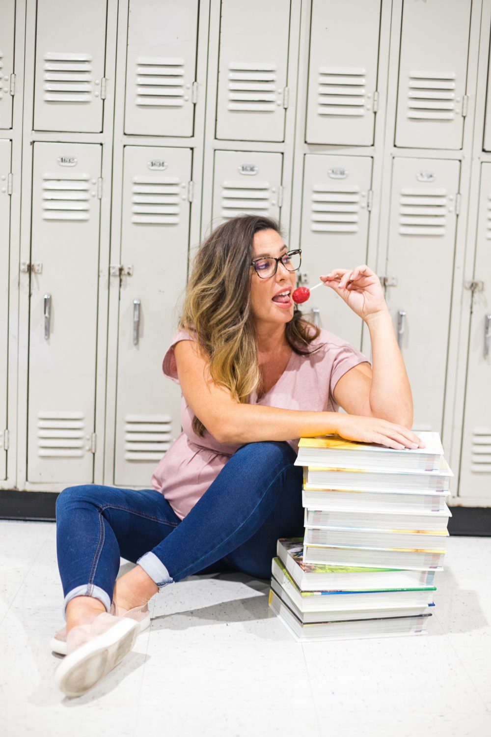 Back to School Eye Exam: How to Prepare your Child for a Successful Academic Year | Back to School Eye Exam: How to Prepare your Child for a Successful Academic Year by popular Florida lifestyle blog, Fresh Mommy Blog: image of a woman sitting in front of some school lockers wearing a pair of blue light glasses, holding a sucker, and resting her elbow on a stack of books. 