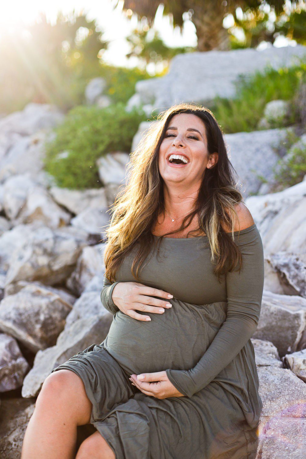 A Beach Maternity Shoot in Green | A Beach Maternity Photoshoot in Olive Green by popular Florida life and style blog, Fresh Mommy: image of a woman sitting outside at the beach and wearing a Pink Blush Olive Solid Off Shoulder Maternity Maxi Dress.