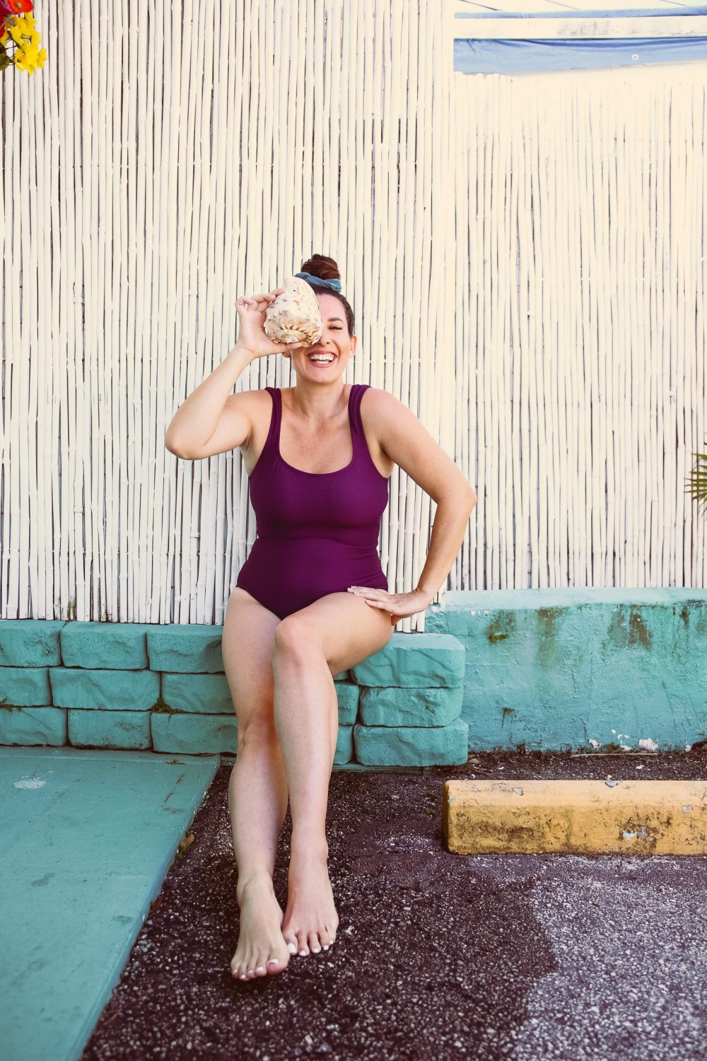 Style on a Budget: 9 Best Swimsuits for Moms |Swimsuits for Moms by popular Florida fashion blog, Fresh Mommy Blog: image of a woman sitting in front of a white bamboo fence and wearing a purple ribbed square neck cap sleeve one piece swimsuit. 